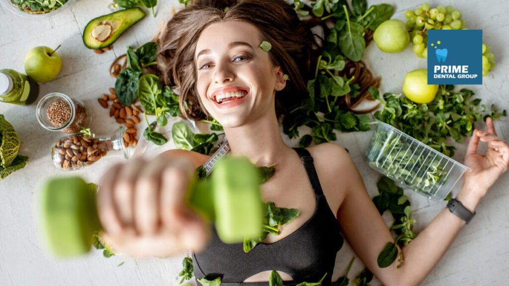 Smiling woman surrounded by healthy, leafy greens and fruits, promoting healthy eating habits for strong teeth and overall wellness, with the Prime Dental Group logo in the corner